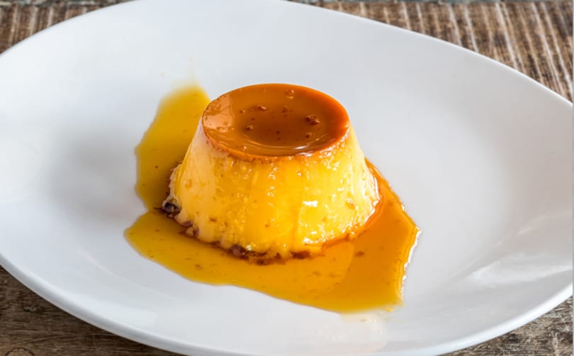 A golden flan dessert with caramel sauce on a white plate, against a rustic wooden background.