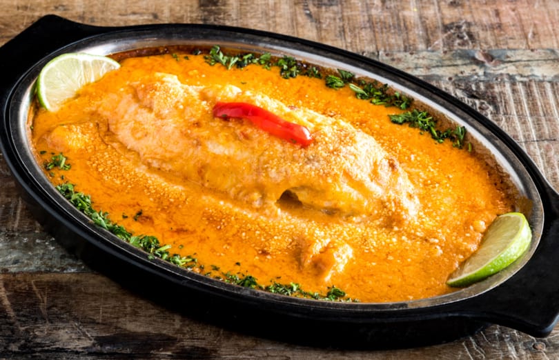 Baked fish in a rich orange sauce, garnished with lime wedges, parsley, and a red pepper.