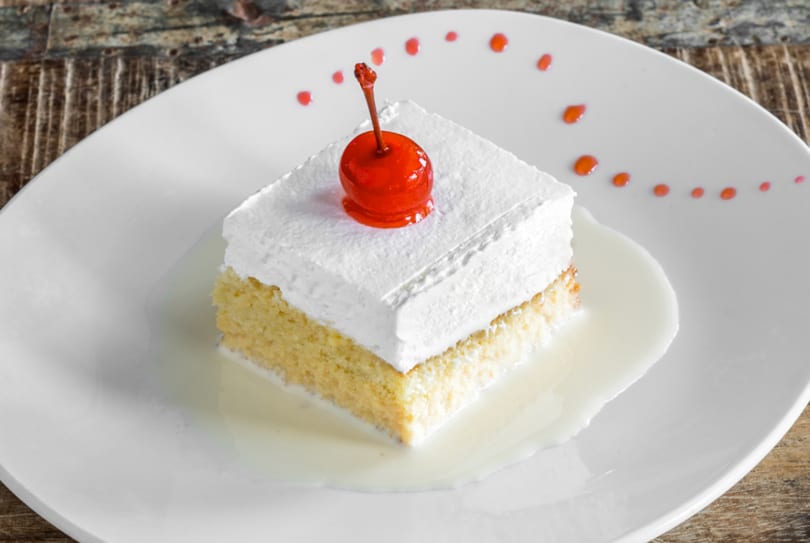A slice of tres leches cake with whipped cream and a cherry on a white plate.