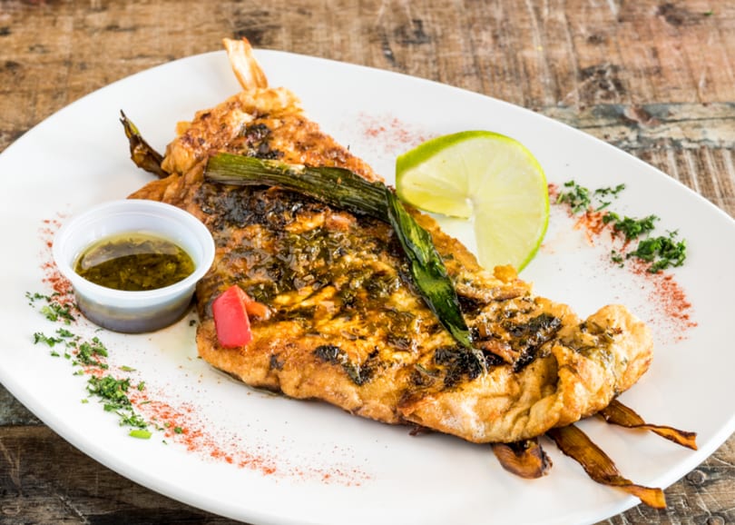 Grilled fish with green onions, lime, and a side of sauce on a white plate.