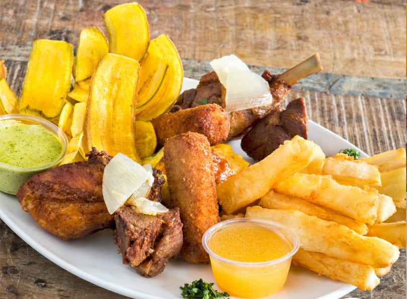 A platter of fried plantains, yuca fries, various meats, and dipping sauces on a wooden table.