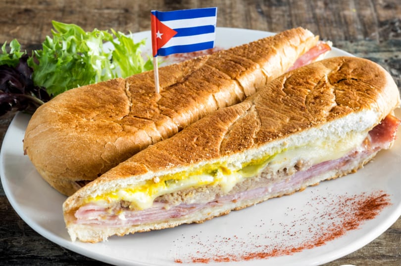 A Cuban sandwich with a Cuban flag toothpick, served with lettuce on a white plate.