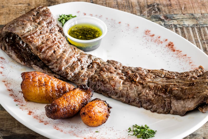 Grilled skirt steak with roasted plantains and a side of chimichurri sauce on a white plate.